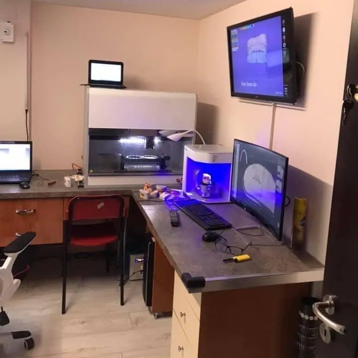 Camera laboratorului: birouri cu monitoare, un scaner dentar luminat în albastru, aparatura de frezare cu ușă de sticlă și un televizor pe perete cu un model 3D al unei arcade.
