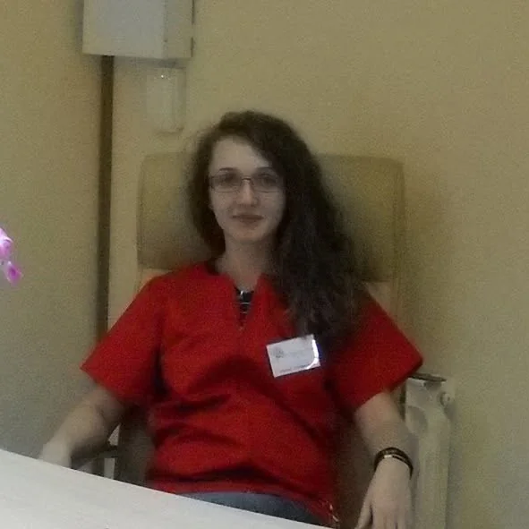 Medicul stomatolog, în uniformă roșie, la biroul din cabinet, lângă o orhidee. Fotografia de profil a paginii de Facebook a cabinetului.