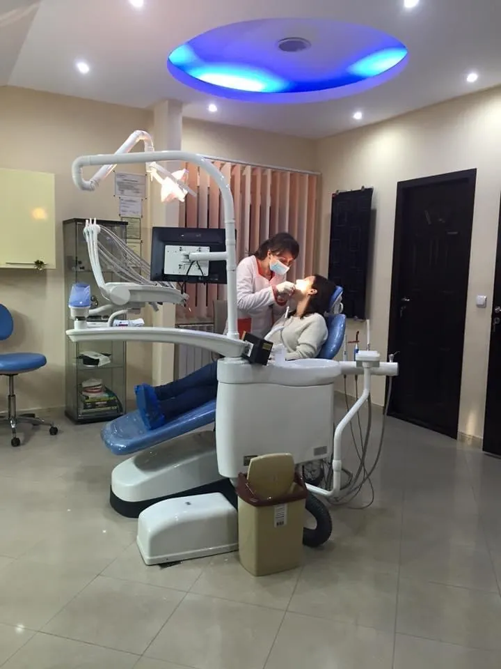 Sala de tratament: fotoliul stomatologic albastru cu alb, lumina albastră din tavan, vitrina cu sertare și o ușă din lemn închis la culoare.