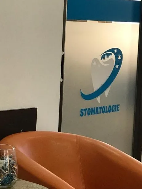 Ușa de sticlă a cabinetului, cu un dinte desenat cu albastru și scrisul „Stomatologie”, lângă un fotoliu portocaliu.